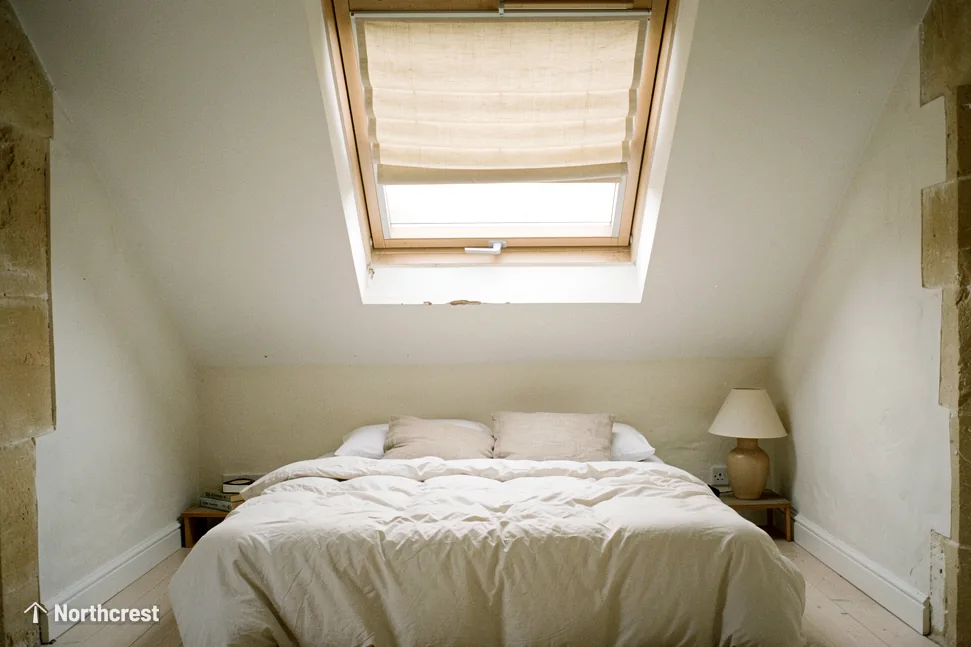 Loft bedroom with stone edged walls under a pine roof window with a roman blind